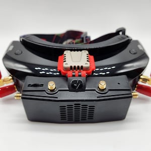 May include: A black and red FPV headset with a built-in fan and two antennas. The headset has a black strap and a silver logo on the front.