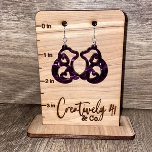 May include: A pair of purple and black marbled acrylic earrings with a unique design, hanging from silver-colored hooks. The earrings are displayed on a wooden stand with the text "Creatively 41 & Co."