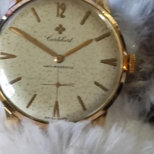 May include: A gold-toned wristwatch with a cream-colored face. The watch features gold hands and markers, with the number 12 prominently displayed. The watch face has the brand name "Carlebert" and the text "ANTI-MAGNETIC" and "SWISS MADE".