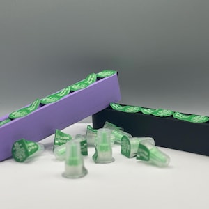 May include: A close-up of a purple and black container with green labels. The containers are filled with small, clear, plastic vials with green labels. The vials are for medical use.