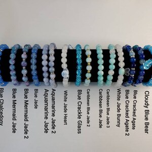 Op de afbeelding: Een verzameling van 15 armbanden gemaakt met verschillende blauwe edelstenen en glazen kralen. De armbanden zijn in een rij op een zwart oppervlak gerangschikt. De namen van elke armband staan ​​onder de armbanden.