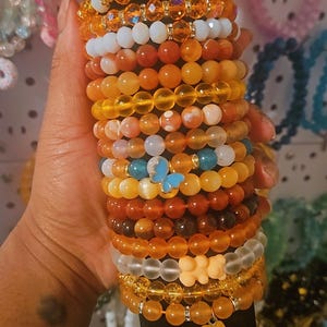 May include: A selection of beaded bracelets in orange, yellow, and brown hues. Some bracelets include clear beads, a blue butterfly charm, and a gummy bear charm. The bracelets are stacked on a black cylindrical display.