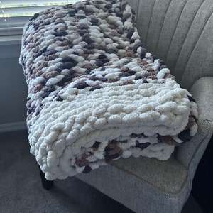 May include: A soft, chunky knit throw blanket in shades of white, brown, and gray. The blanket is folded and draped over a gray armchair.