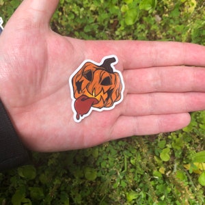 May include: A sticker of a jack-o'-lantern with a wide, toothy grin and a long, red tongue sticking out. The pumpkin is orange and has a green stem.