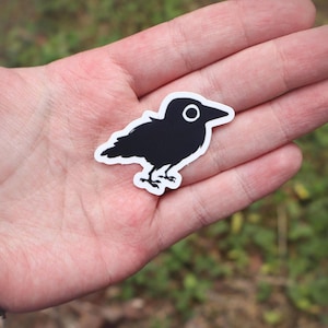May include: A black and white sticker of a crow with a white outline. The crow is in a simple, cartoon style, with a round eye and detailed feet. The sticker is held in a hand against a blurred green background.