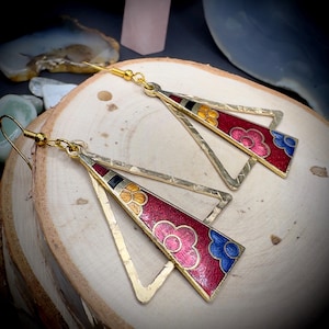 May include: A pair of gold-tone earrings with a triangular design. The earrings feature a red enamel inlay with floral patterns in pink, yellow, and blue. The earrings have a hammered texture and a hook closure.