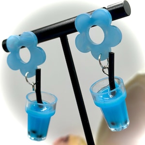 May include: Light blue flower-shaped earrings with a clear plastic cup of blue liquid and black boba pearls. Each earring has a black straw. The earrings are displayed on a black stand.