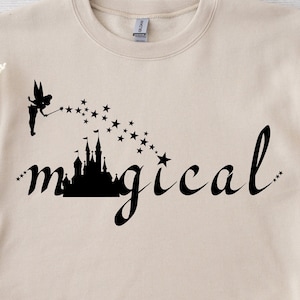 May include: A beige sweatshirt with a black graphic of a fairy flying over a castle with stars and the word "magical" written in cursive.