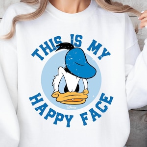 May include: White sweatshirt featuring a cartoon image of Donald Duck with the text "THIS IS MY HAPPY FACE" in blue. The graphic includes Donald Duck's face inside a light blue circle.