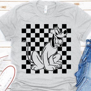 Könnte beinhalten: Hellgraues T-Shirt mit einem schwarz-weißen Schachbrettmuster und einer Cartoon-Illustration von Pluto. Der Cartoon-Hund sitzt und lächelt. Weiße Turnschuhe und blaue Jeans sind sichtbar.