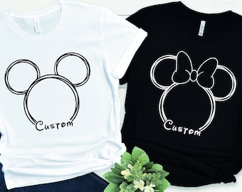 Op de afbeelding: Twee T-shirts met een witte omtrek van de hoofden van Mickey Mouse en Minnie Mouse. Het Mickey Mouse shirt is wit en het Minnie Mouse shirt is zwart. Beide shirts hebben het woord "Custom" onder de omtrek gedrukt.