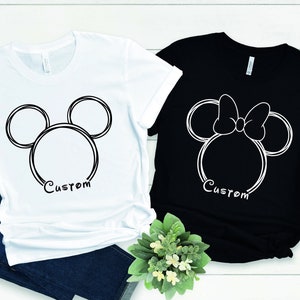 May include: Two t-shirts with a white outline of Mickey Mouse and Minnie Mouse heads. The Mickey Mouse shirt is white and the Minnie Mouse shirt is black. Both shirts have the word "Custom" printed below the outline.