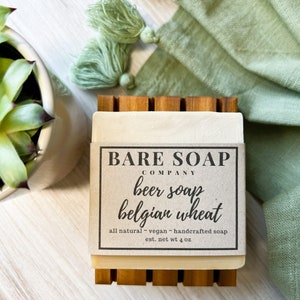 May include: A bar of natural beer soap with a brown paper label that reads "BARE SOAP COMPANY beer soap belgian wheat all natural vegan handcrafted soap est. net wt 4 oz". The soap is sitting on a wooden soap dish.