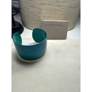 May include: A teal blue metal bangle bracelet. The bracelet is wide and has a rounded shape. The bracelet is sitting on a white surface. The text "neon brights bracelet" is visible on a small white box.
