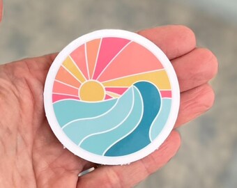 May include: A round sticker with a colorful sunset and a blue wave design. The sticker has a white border and is perfect for adding a touch of summer to your belongings.