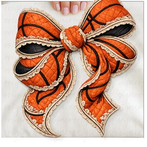 May include: An orange and black bow with a basketball design. The bow is made of quilted fabric and features black lines, resembling the seams of a basketball. The edges are trimmed with a cream-colored lace.