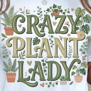 Puede incluir: Camiseta blanca con el texto "CRAZY PLANT LADY" en letras verdes y beige. El diseño incluye ilustraciones de plantas en macetas, hojas y corazones, creando un tema botánico.