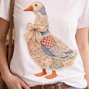 Può includere: T-shirt bianco con un design a patchwork di un'oca. L'oca ha un fiocco beige, zampe arancioni e un collo ornato di pizzo. Il patchwork include motivi floreali, a quadretti e a pois. La maglietta è abbinata a jeans blu e una cintura marrone.