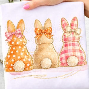 Könnte beinhalten: Auf weißem Hintergrund sind drei Aquarell-Hasenillustrationen zu sehen. Jeder Hase wird von hinten betrachtet, mit flauschigen weißen Schwänzen und goldenen Glitzerakzenten. Die Hasen tragen Schleifen in rosa Vichy, orangefarbenen Punkten und orangefarbenen Punkten.