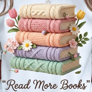 Könnte beinhalten: Weißes T-Shirt mit einem Design aus einem Stapel gehäkelter Bücher in Pastellfarben, verziert mit gehäkelten Blumen und dem Text "Read More Books". Die Bücher sind in Creme, Rosa, Pfirsich, Lavendel und Mintgrün.