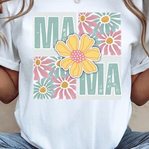 May include: White t-shirt with a floral design and the word "MAMA" in green and white letters.