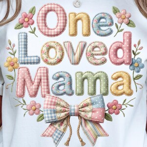 Può includere: T-shirt bianco con la scritta "One Loved Mama" in lettere colorate in stile patchwork. Il design include elementi floreali e un fiocco, creando un'estetica affascinante e femminile.