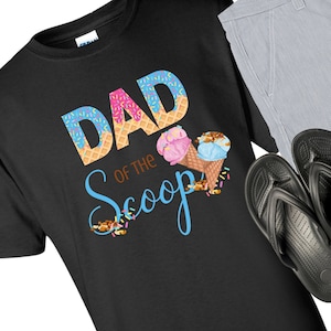 May include: Black t-shirt with the text "Dad of the Scoop" in blue and brown lettering. The text is surrounded by ice cream cones with pink and blue ice cream, sprinkles, and nuts.
