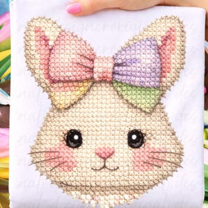 May include: A cross-stitch design of a bunny face with a colorful bow. The bunny has black eyes, pink cheeks, and a pink nose. The bow features pink, purple, and green sections. The design is on a white background.
