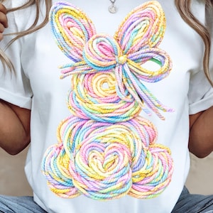 Puede incluir: Camiseta blanca con un diseño de conejito colorido. El conejito está hecho de cuerda enrollada en tonos pastel de rosa, amarillo, azul y verde, con un detalle de lazo. El diseño está centrado en la camiseta.