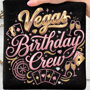 Könnte beinhalten: Schwarzes T-Shirt mit einem goldenen und rosa Design. Das Design enthält die Worte "Vegas Birthday Crew", Spielkarten, Sektgläser und ein Rouletterad. Das T-Shirt wird von einer Hand gehalten.