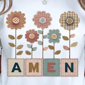 Könnte beinhalten: Weißes T-Shirt mit einem Blumendesign und dem Wort "AMEN". Die Blumen sind in Braun-, Rosa- und Blautönen gehalten, mit detaillierten Stielen und Blättern. Die Buchstaben sind im Patchwork-Stil gestaltet.