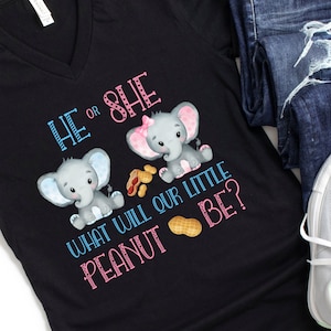Puede incluir: Camiseta negra con cuello en V con el texto "HE or SHE WHAT WILL OUR LITTLE PEANUT BE?" en azul y rosa. La camiseta presenta ilustraciones de dos elefantes grises con orejas rosas y azules y cacahuetes.