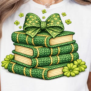 May include: A white t-shirt featuring a stack of green books with yellow accents, topped with a green bow. The books and bow have a knitted texture. Green shamrocks and flowers are scattered around the books.