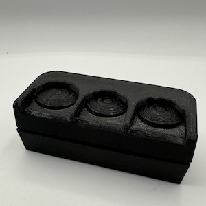 Op de afbeelding: Een zwarte, rechthoekige 3D-geprinte doos met drie cirkelvormige inkepingen. De inkepingen hebben een spiraalvormig ontwerp. De doos is gemaakt van een matzwart materiaal en heeft een scharnierend deksel.