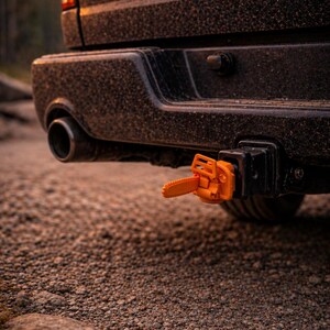 Könnte beinhalten: Nahaufnahme einer schwarzen LKW-Stoßstange mit einer orangefarbenen Anhängerkupplungsabdeckung in Form einer Kettensäge. Der LKW ist mit Schmutz bedeckt, vor einem Schotterweg und Bäumen.