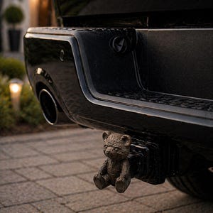 May include: A bronze teddy bear hitch cover fixed to the back of a black truck. The bear is depicted sitting with its arms crossed. The truck's bumper and exhaust pipe are also visible.