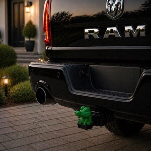 May include: Black RAM truck rear with chrome lettering and a green alligator hitch cover. The truck is parked on a brick driveway in front of a house with a black door and outdoor lighting.