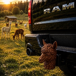 Op de afbeelding: Een bruine, gebeeldhouwde alpaca-kop trekhaakdop bevestigd aan de achterbumper van een zwarte RAM-truck. De trekhaakdop heeft een gedetailleerde vachtstructuur en puntige oren, geplaatst op de trekhaakontvanger van het voertuig.