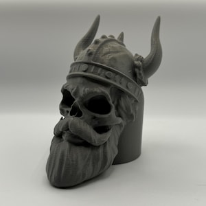 Peut inclure: Un crâne gris imprimé en 3D portant un casque à cornes. Le crâne a une longue barbe et un visage détaillé.