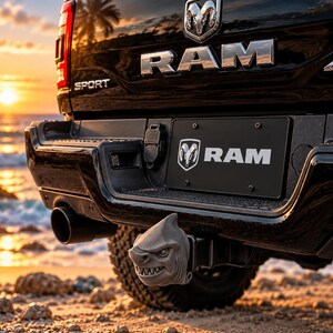 Op de afbeelding: Een grijze haai-vormige trekhaakdop gemonteerd op de achterbumper van een zwarte RAM Sport pick-up. Het voertuig is voorzien van verchroomde RAM-belettering, een 4x4-embleem en een zwarte kentekenplaat met het RAM-logo en de tekst.