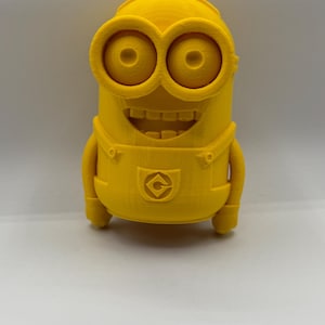 Op de afbeelding: Een geel 3D-geprint figuurtje van een minion uit de Despicable Me-films. De minion heeft een grote glimlach, grote ogen en een G op zijn borst.