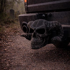 May include: Black skull hitch cover with horns attached to a black truck bumper. The word "RAM" is visible on the truck. The skull has detailed features and is set against a blurred outdoor background.