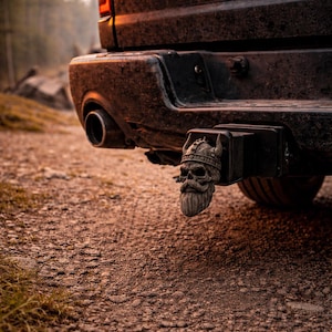 Può includere: Primo piano della parte posteriore di un camion nero, con una copertura decorativa per gancio di traino a forma di teschio vichingo con barba e casco con corna. Il camion è parcheggiato su una strada sterrata, con la parola "RAM" visibile.