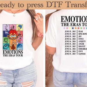 以下が含まれることがあります： 内側にアウトのキャラクターのグラフィックが描かれた白いTシャツ。グラフィックの下に「Emotions The Eras Tour」というテキストが印刷されています。シャツの背面には同じテキストと日付と感情のリストがあります。