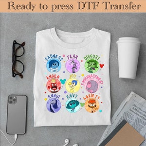 以下が含まれることがあります： 内側にアウトのキャラクターのカラフルなグラフィックが描かれた白いTシャツ。キャラクターは名前が書かれた円形に配置されています。名前は、悲しみ、恐怖、嫌悪、怒り、喜び、恥ずかしさ、退屈、嫉妬、不安です。