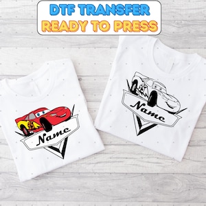 Può includere: Due magliette bianche con un design di auto da corsa di cartoni animati rosso e nero. Il design include le parole "Nome" in uno stendardo. Le magliette sono piegate e mostrate su una superficie di legno. Trasferimento DTF pronto per la stampa.