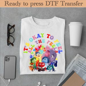 以下が含まれることがあります： 白いTシャツに、映画「インサイド・ヘッド」のキャラクターがカラフルなグラフィックで描かれています。グラフィックには「すべての感情を感じることは大丈夫」と書かれています。