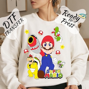 Op de afbeelding: Witte sweatshirt met een kleurrijke Super Mario-afbeelding. Het ontwerp bevat Mario, Bowser en andere personages, met de tekst "Super Mario". De woorden "DTF TRANSFER" en "Ready to Press" zijn ook zichtbaar.