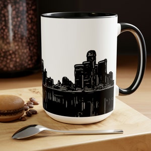 Könnte beinhalten: Eine weiße Keramiktasse mit schwarzem Rand und Henkel. Die Tasse zeigt eine schwarze Silhouette einer Stadtlandschaft.