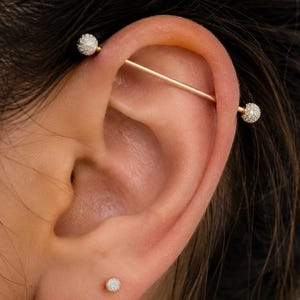 May include: A gold-toned industrial barbell piercing with two spherical ends covered in small clear crystals, positioned through the upper cartilage of an ear. A small, single stud earring with a clear crystal is located in the earlobe below.